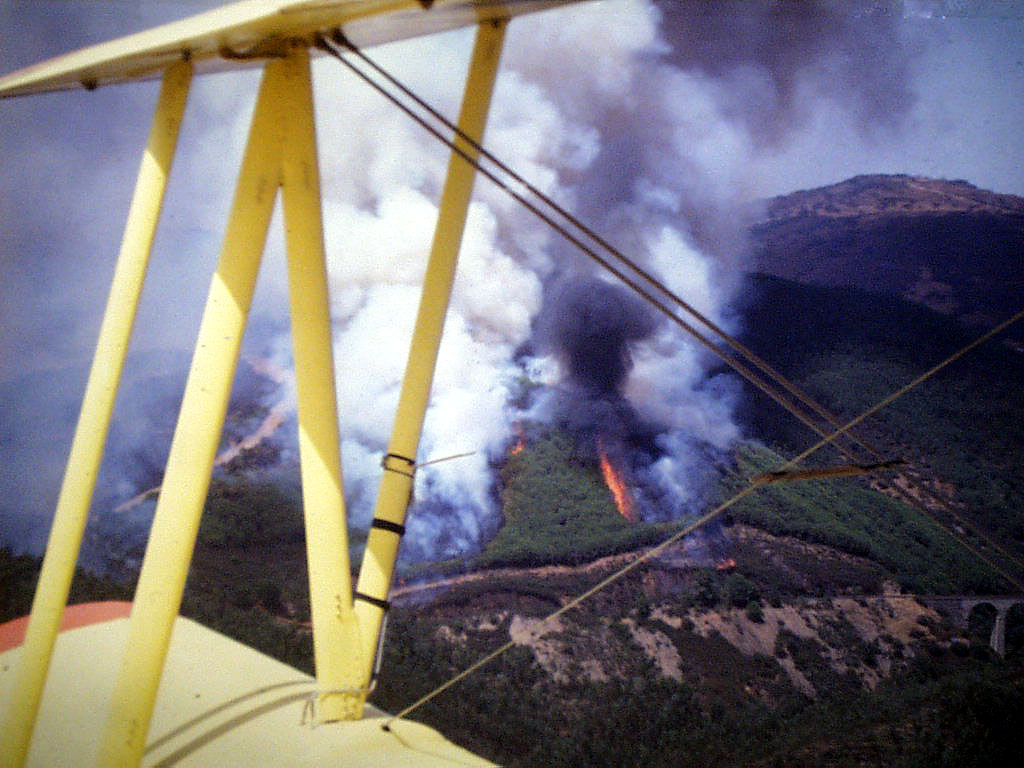 Incendio en ladera con humo negro visto desde el ala del biplano amarillo; al fondo, un viaducto